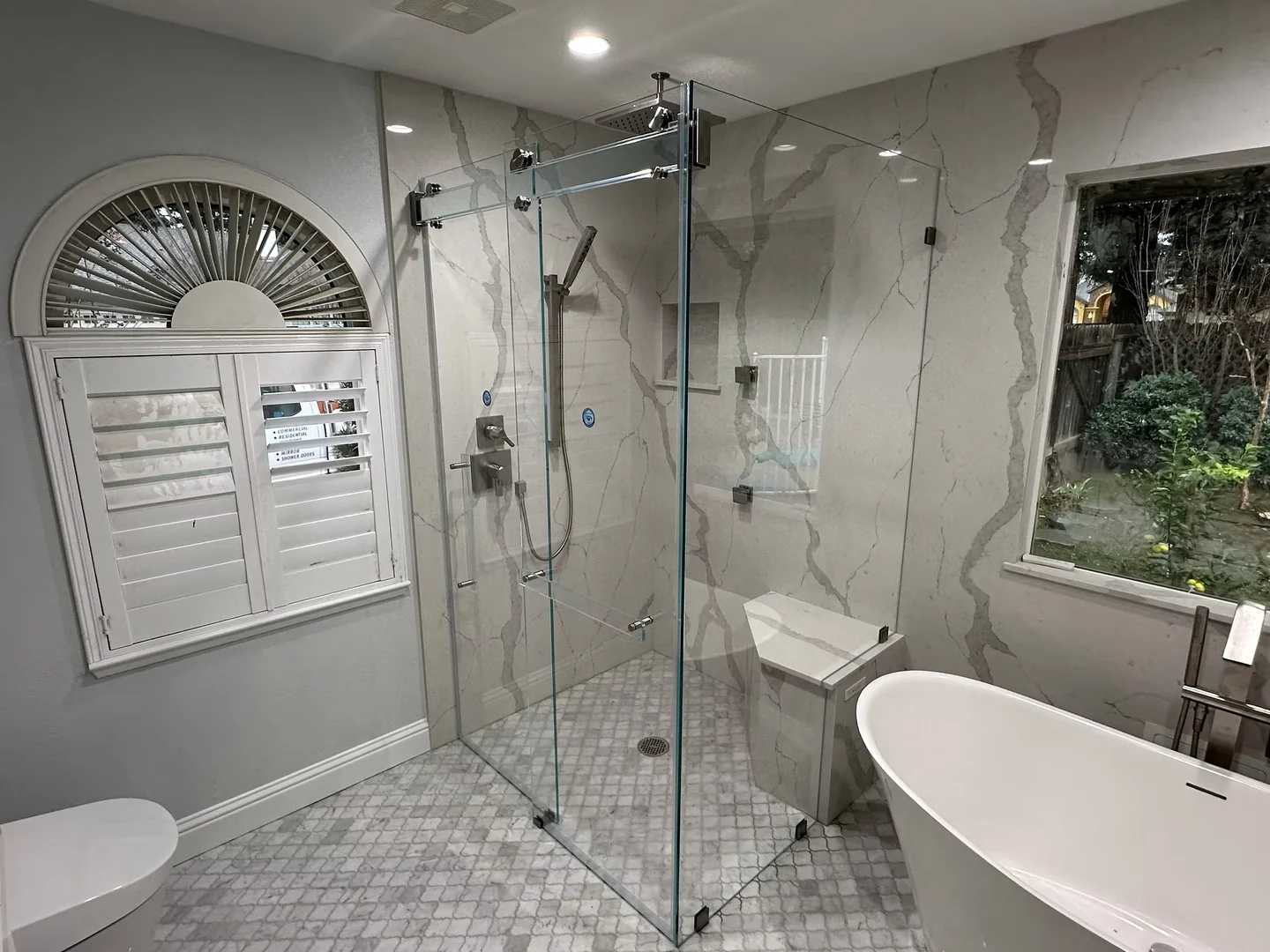 Titan Phantom corner shower with a glass header and polished fittings beside an arched window and freestanding tub.