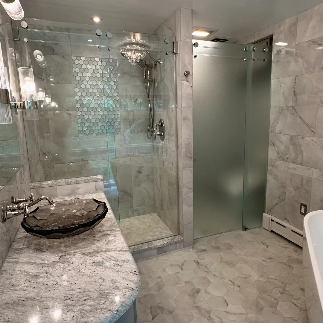 Clear glass-header shower with polished fittings alongside a separate frosted glass enclosure in a marble bathroom.