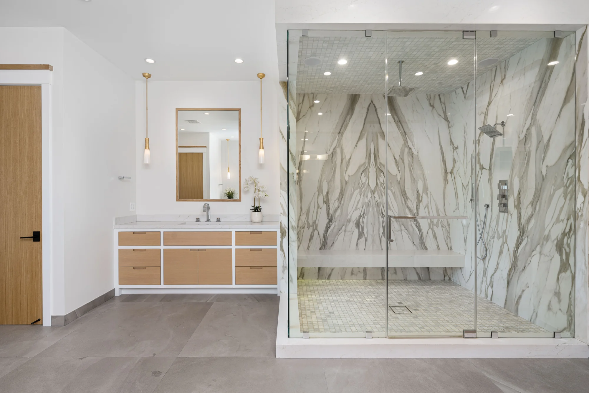Clear frameless shower enclosure beside a wood vanity, with a bench and pale veined walls.