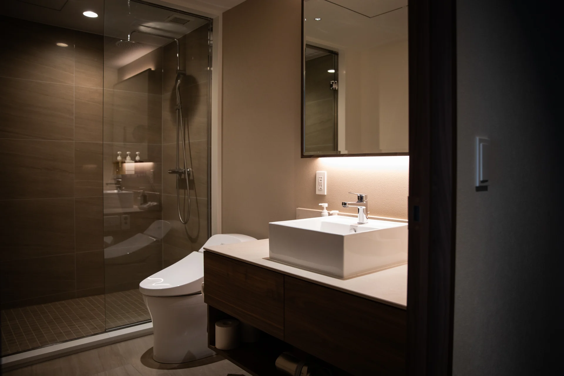 Clear glass shower enclosure against warm dark tile in a softly lit bathroom.