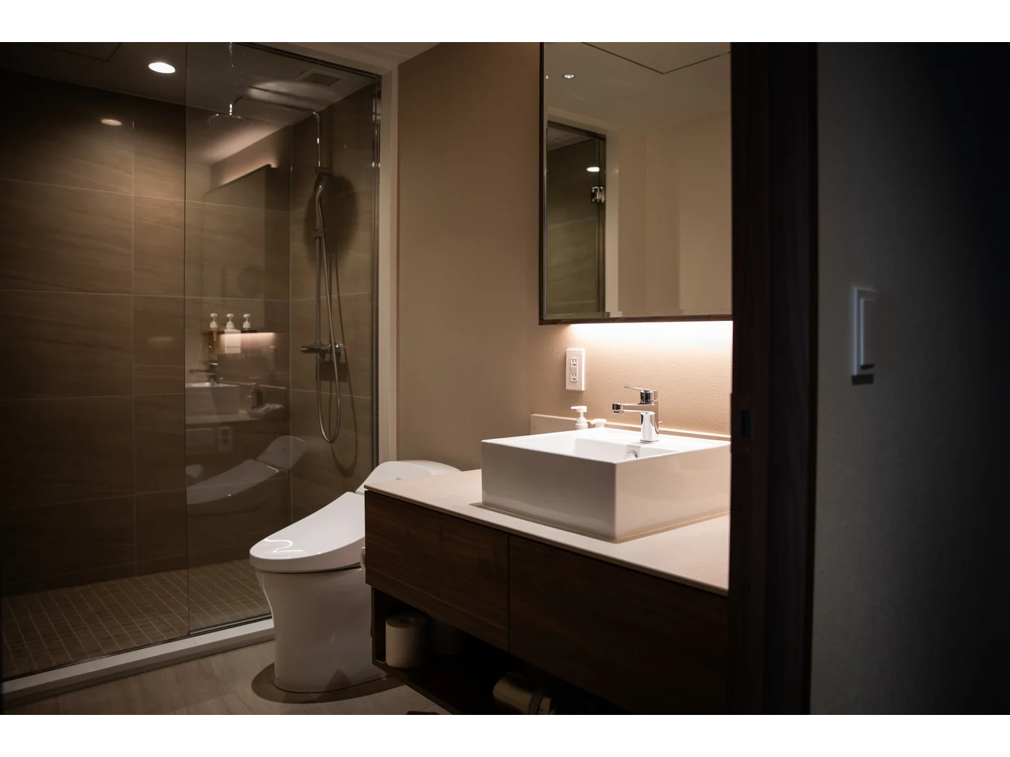 Clear glass shower enclosure against warm dark tile in a softly lit bathroom.