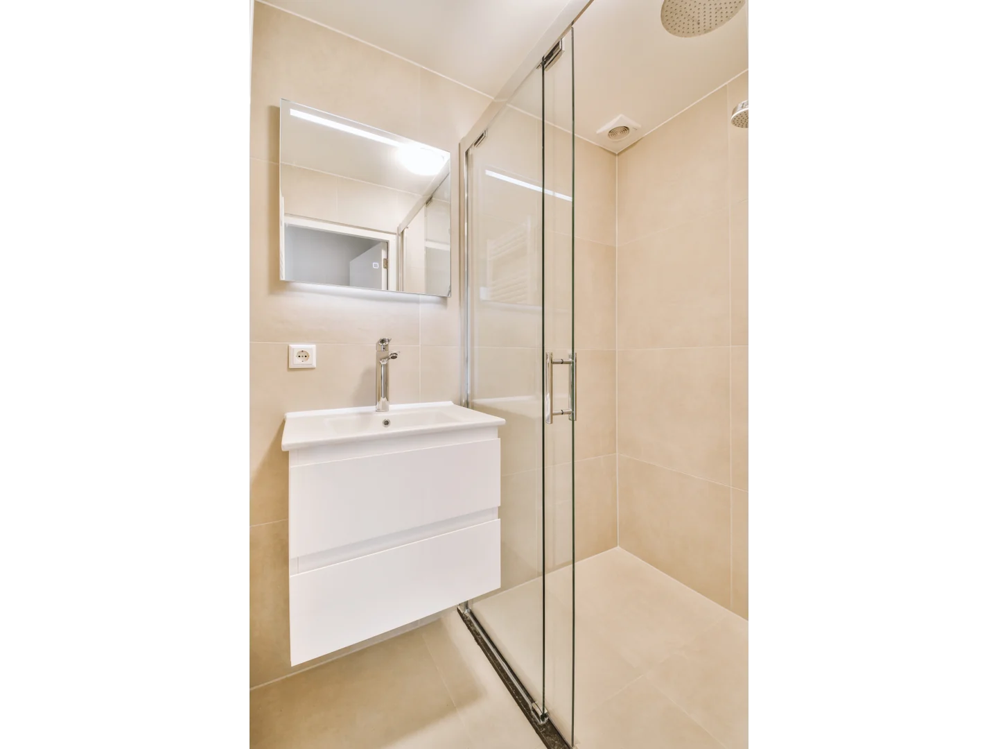 Clear glass shower panels with slim silver-colored profiles beside a white vanity in a pale bathroom.
